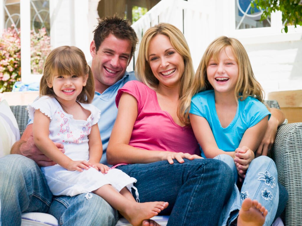 A smiling mother, father, and their two young daughters sitting closely together on a sofa, optimized as a Family & Parenting image on home page of beautyrelationship.com.