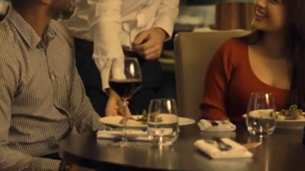 A couple smiling at an upscale restaurant table while a waiter serves food, showcasing classic and elegant budget friendly dinner date night outfits.