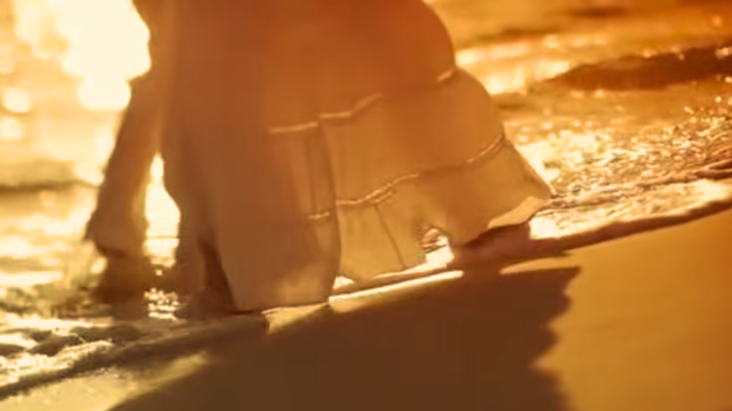 A close-up shot of a couple's lower half as they walk barefoot along the seashore at sunset, with the woman wearing a flowy white tiered linen maxi skirt that lightly brushes against the incoming ocean waves on the wet sand.